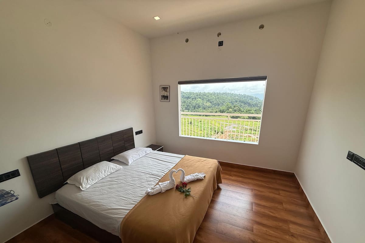 A bedroom with a large window, wooden flooring, a bed with white linens, and a brown throw blanket.