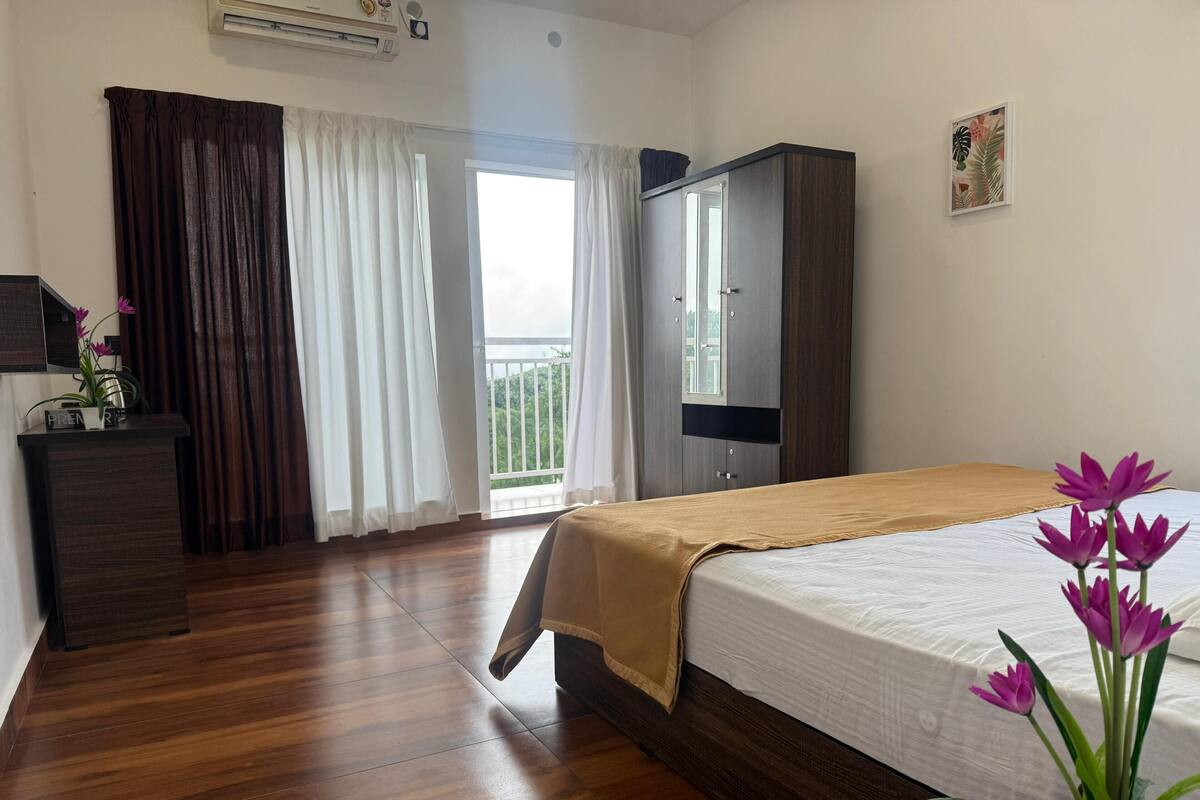 A hotel room with a wooden floor, a bed with a beige bedspread, a desk with a nameplate, a wardrobe, and a window with curtains.