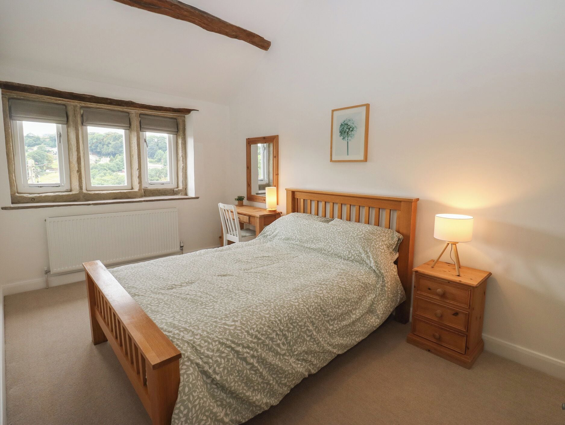 Une chambre avec un lit en bois, une table de chevet avec une lampe, un bureau avec une chaise, un radiateur et une fenêtre donnant sur un paysage verdoyant.