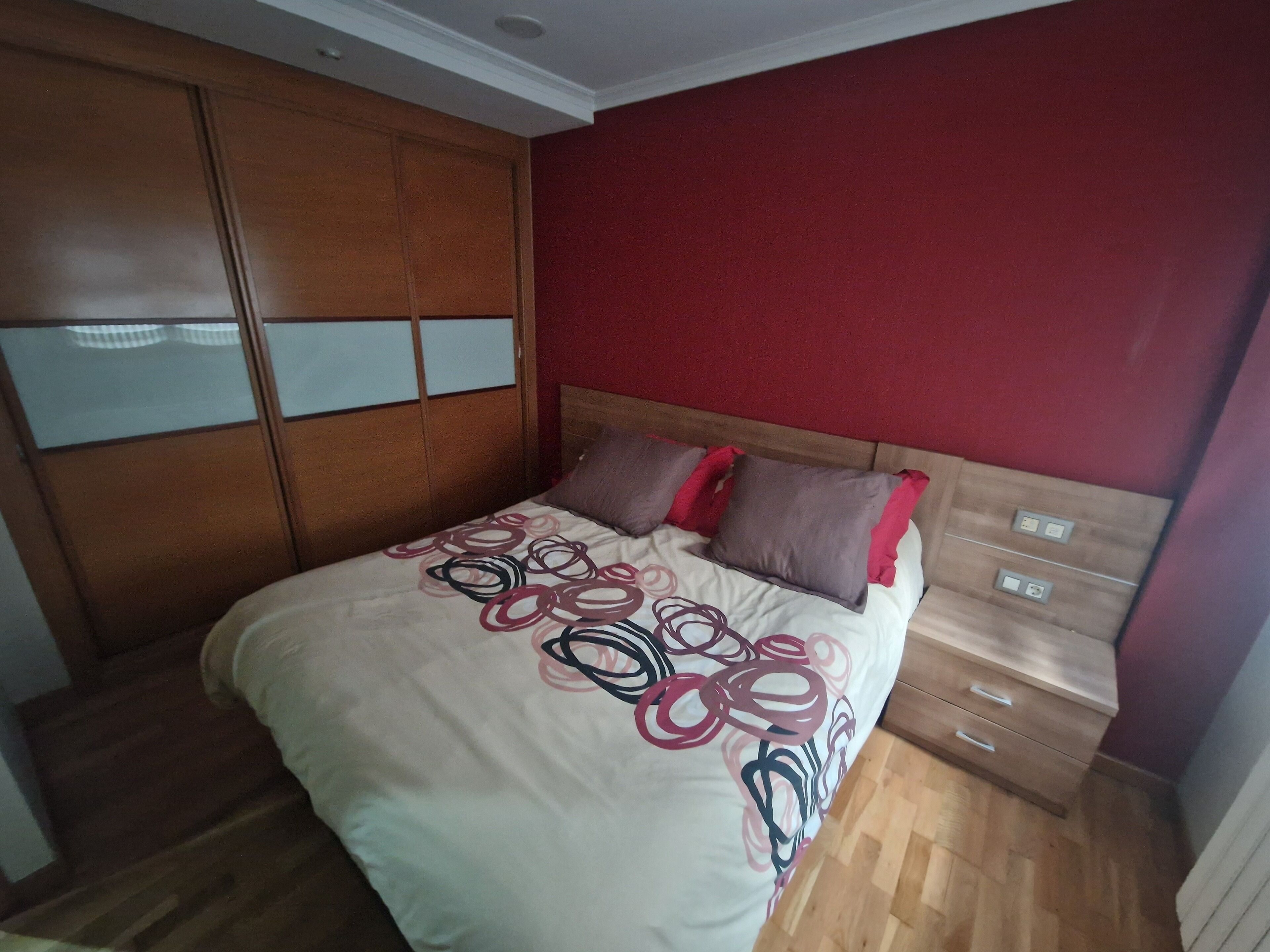 A bedroom with a bed, wooden wardrobe, and a red wall.