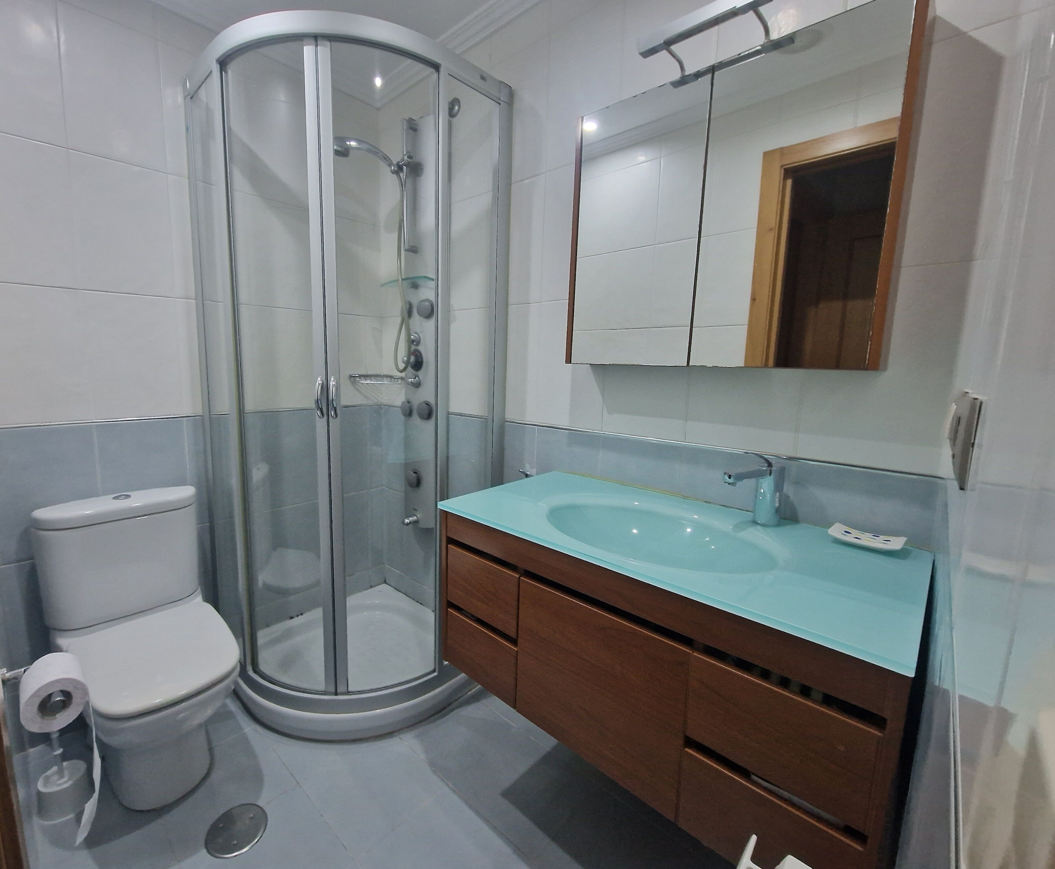 Een moderne badkamer met een douchecabine van glas, een wit toilet en een houten wastafelmeubel met een blauwe wasbak.