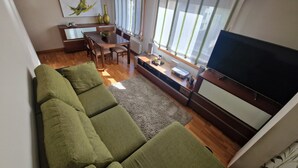 Uma sala de estar com sofá verde, uma mesa de jantar com cadeiras, uma TV e uma janela com cortinas.