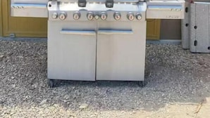Una parrilla de gas portátil con varios quemadores y un quemador lateral, ubicada al aire libre frente a un edificio con puerta amarilla.