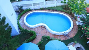 A swimming pool with a brick border, surrounded by greenery and a white fence. - Anton White Villas Resort (Mahabalipuram)