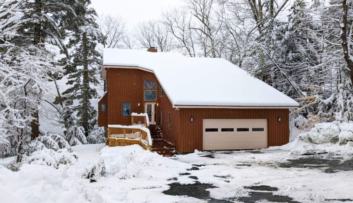 A wooden cabin with a snow-covered roof, a garage, and a small porch.