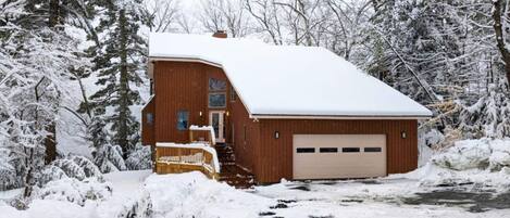 A wooden cabin with a snow-covered roof, a garage, and a small porch.