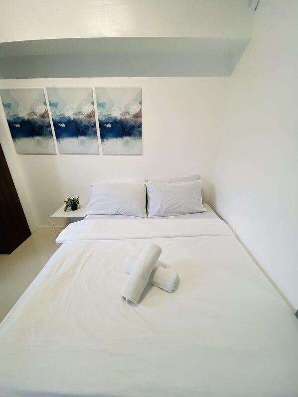 A neatly made bed with white linens and a rolled towel, a small bedside table with a plant, and three abstract paintings on the wall.