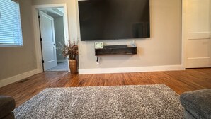 A living room with a flat-screen TV mounted on the wall, wooden flooring, a gray area rug, and a potted plant. - The Carriage House in East Lawrence (Lawrence)