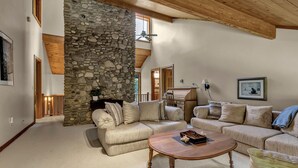 A spacious living room with a stone wall, wooden ceiling beams, and a large window. - Secluded EVL Getaway with Hot tub, Sauna, Fire Pit, Gym & Game room (Ellicottville)