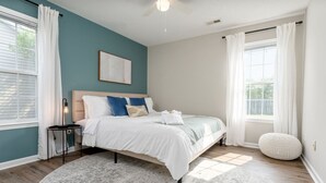 A bedroom with a bed, a bedside table, a lamp, a window with curtains, a wall art piece, and a round ottoman.