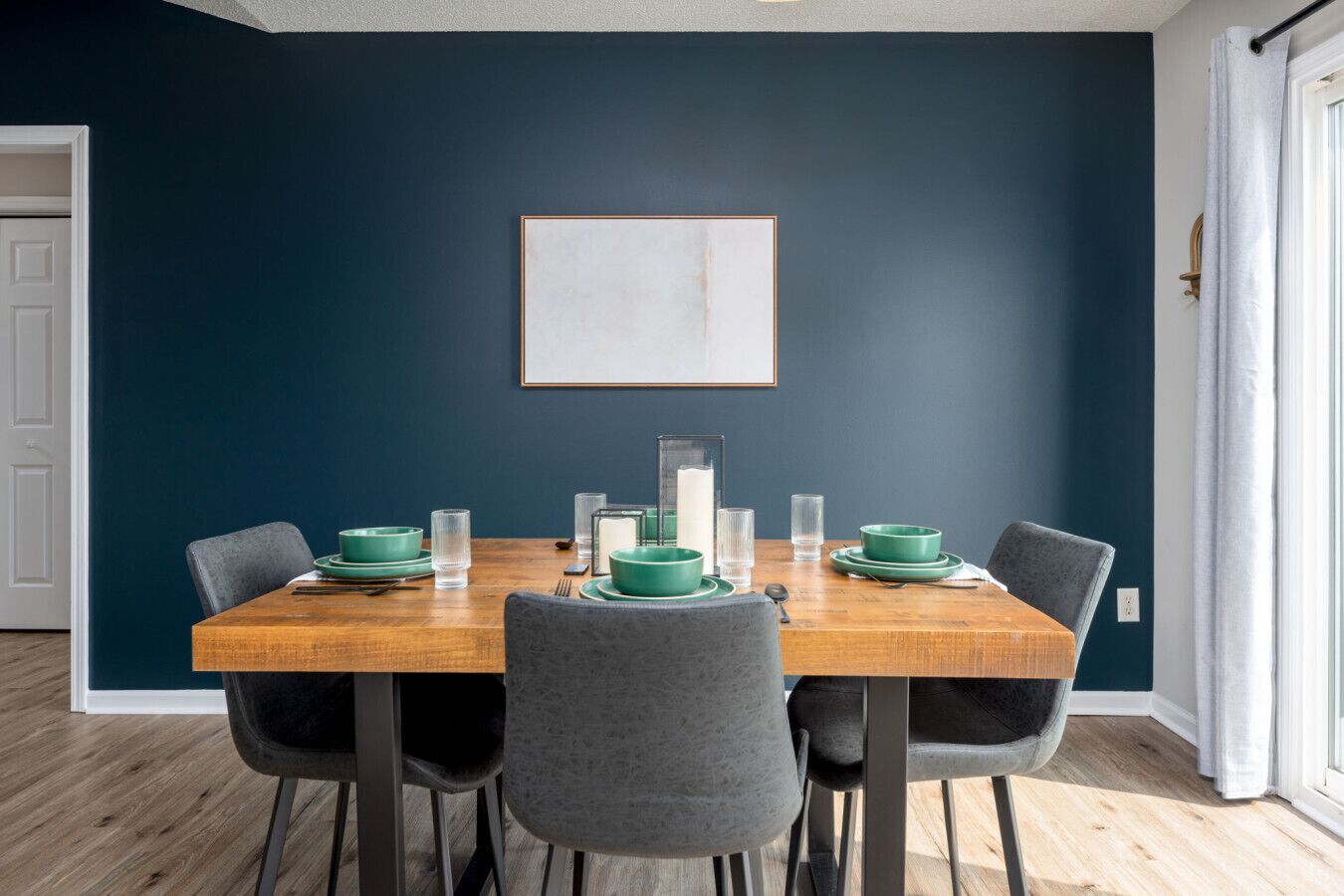 Ein Essbereich mit einem Holztisch, gedeckt für vier Personen, umgeben von grauen Stühlen. Über dem Tisch hängt ein gerahmtes Kunstwerk an der dunkelblauen Wand.