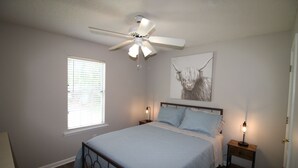 A bedroom with a bed, a ceiling fan, a window with blinds, a bedside table with a lamp, and a wall-mounted artwork. - Simple Stay at Spring Meadow (Valdosta)