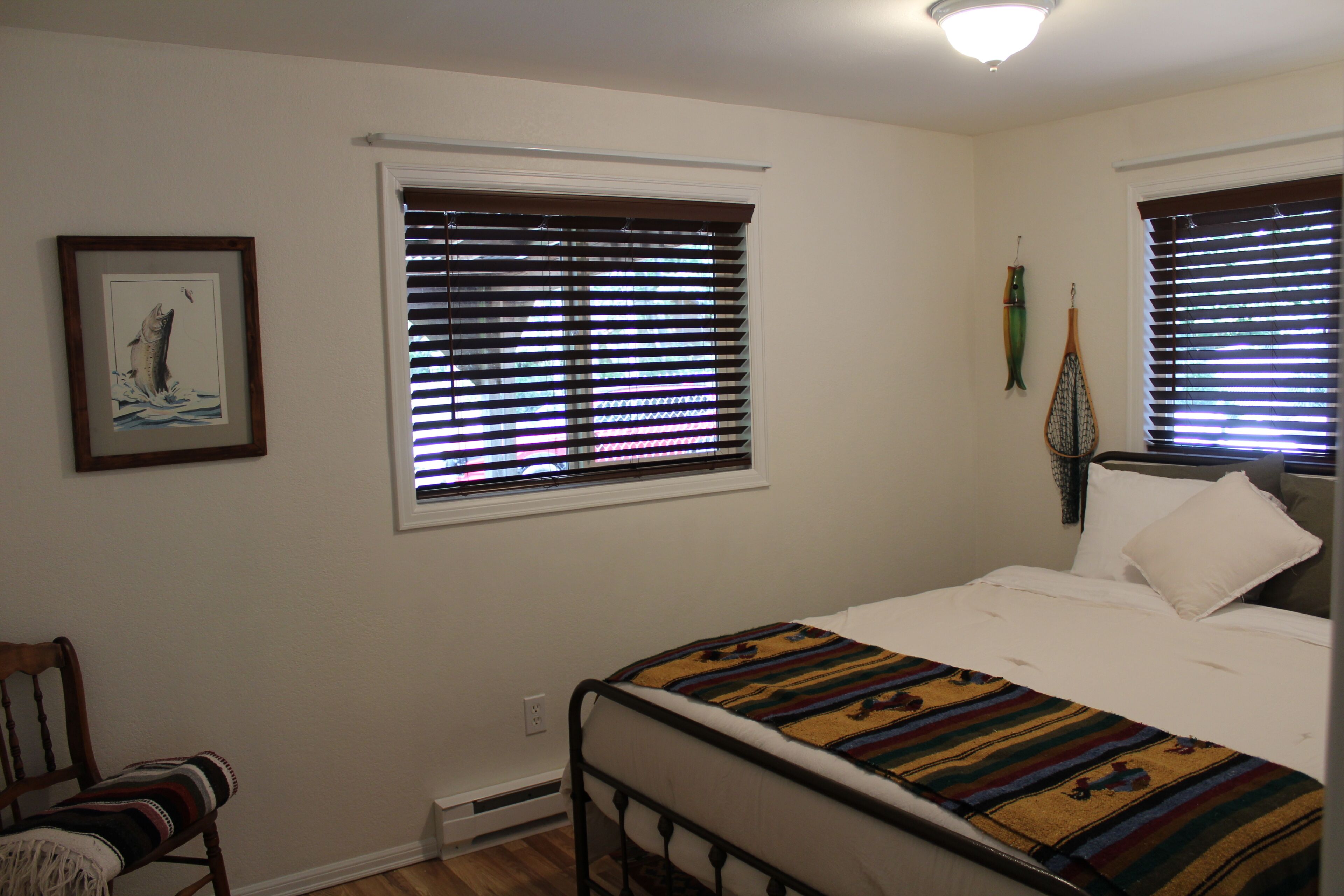 Un dormitorio con una cama, dos ventanas con persianas, un cuadro en la pared y una silla con una manta.