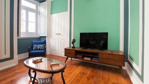 Een kamer met een houten vloer, een glazen salontafel, een televisie op een houten dressoir, een blauwe stoel en een raam met witte luiken.