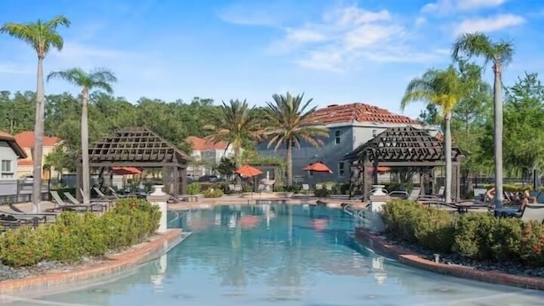 A pool area with lounge chairs, palm trees, and a building with a red roof. - Bella Vida Resort Home in Kissimmee (Kissimmee)