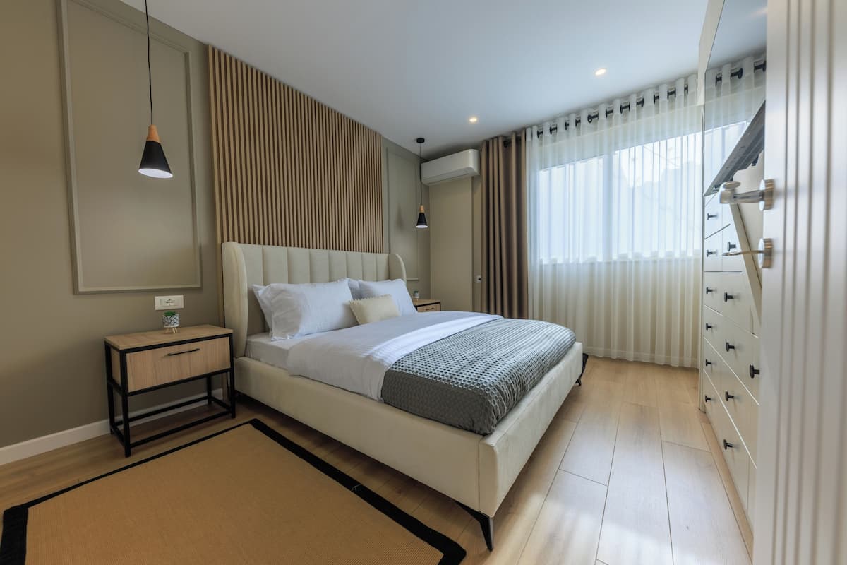 A modern bedroom with a bed, bedside tables, a nightstand, a dresser, and a window with curtains.