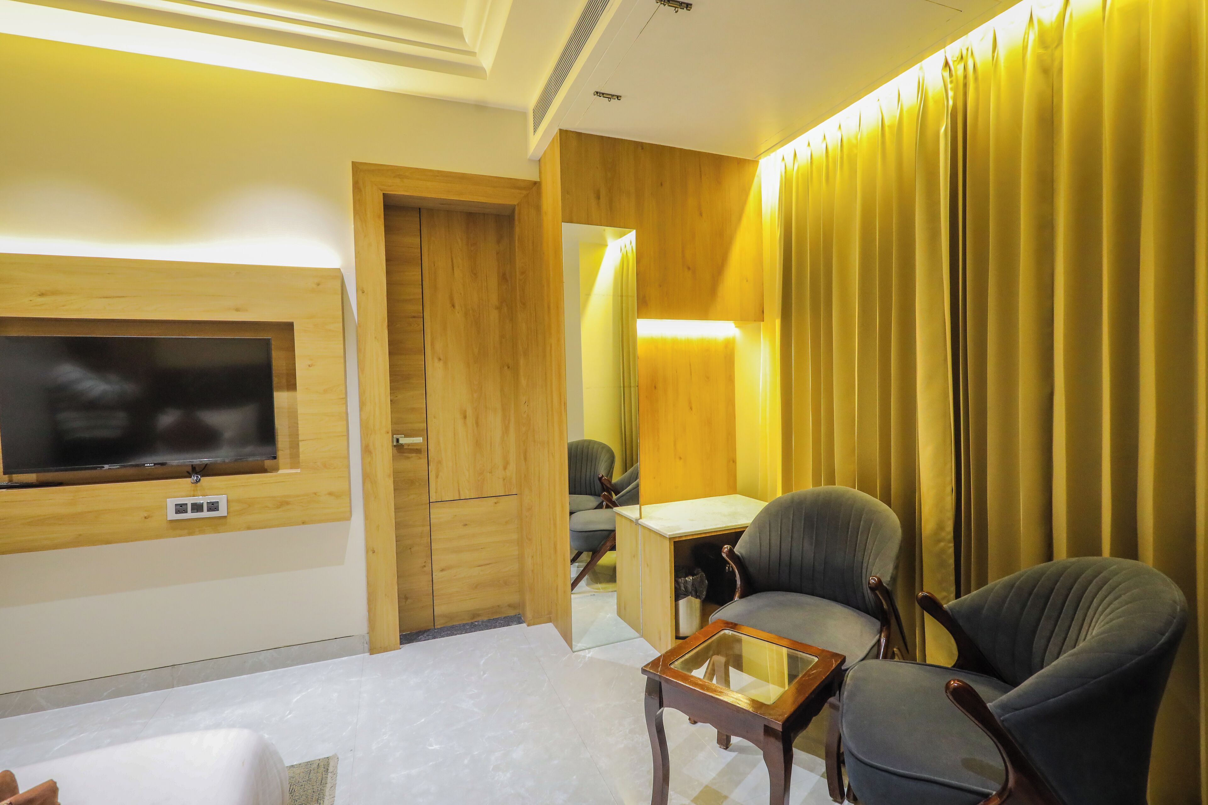 A hotel room with a wooden wall, a flat-screen TV mounted above a wooden cabinet, a desk with a chair, and a large window with yellow curtains.