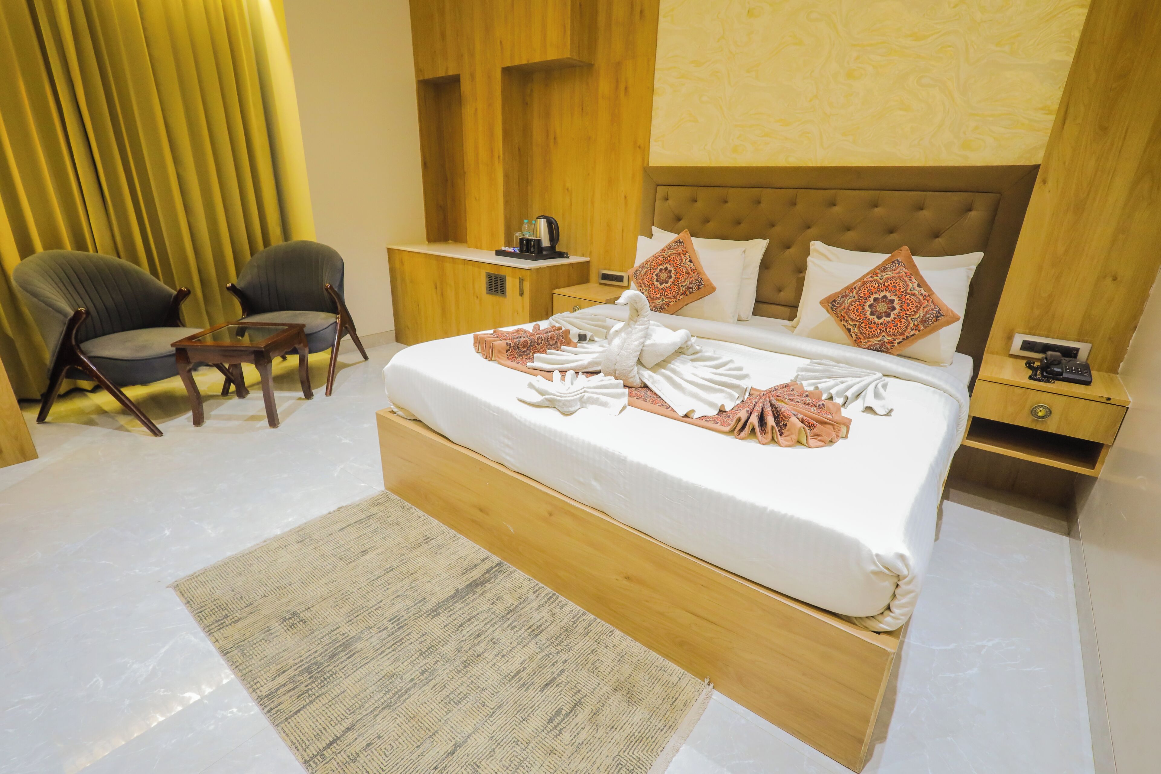 A hotel room with a large bed, two armchairs, a small table, a nightstand, and a wooden headboard.