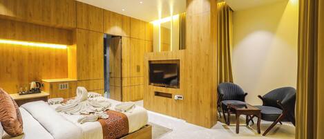 A modern bedroom with a wooden headboard, a flat-screen TV, a bedside table with a lamp, and two chairs.