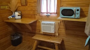 L’intérieur d’une cabane en bois, avec un four à micro-ondes, une petite fenêtre avec des rideaux, une étagère en bois sur laquelle se trouve un plumeau, et une poubelle.