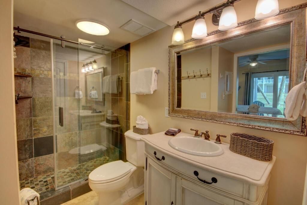 Un baño con ducha de vidrio, inodoro blanco, tocador con lavabo y un espejo grande.