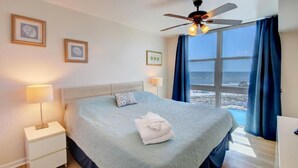 A hotel room with a bed, a nightstand, a ceiling fan, and a large window with a view of the sea.