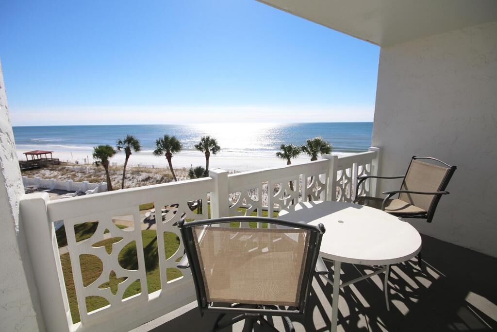 Un balcon doté d’une balustrade blanche, de deux chaises et d’une table, offrant une vue sur une plage bordée de palmiers et sur l’océan.