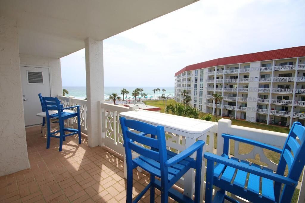Un balcón con sillas azules y una mesa blanca con vista a una playa y un edificio de hotel.