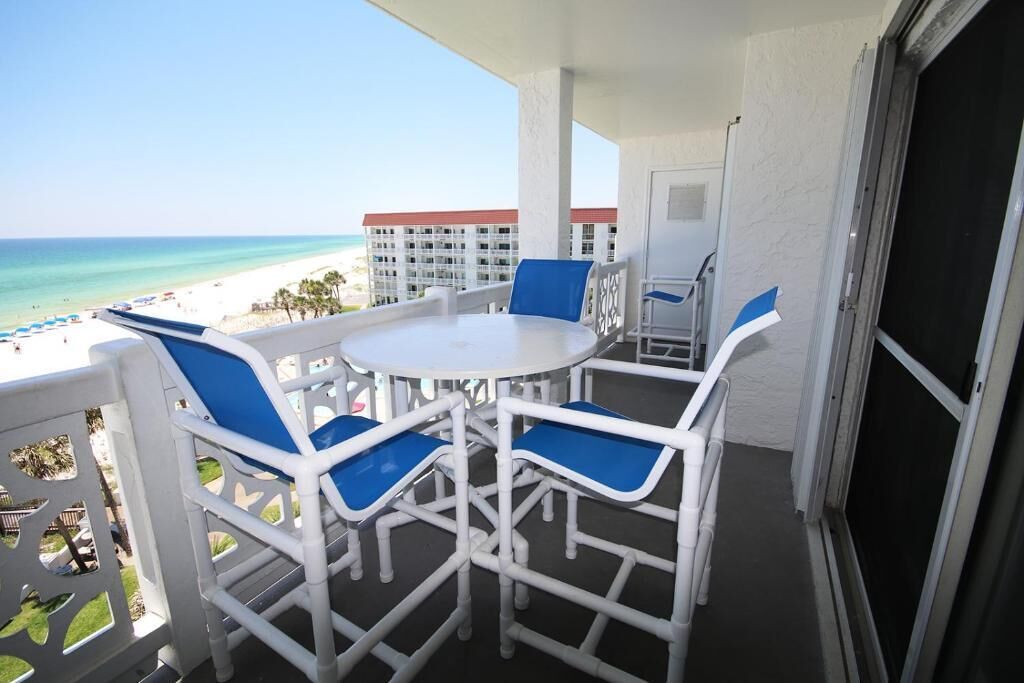 A balcony with a white table and blue chairs, overlooking a beach and hotel buildings.