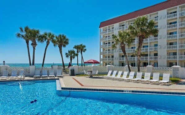 A hotel pool area with a clear view of the ocean, palm trees, and a building with balconies. - El Matador (Fort Walton Beach)
