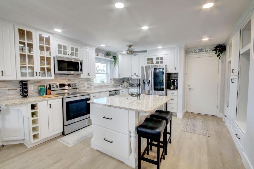 A modern kitchen with white cabinetry, a central island with stools, stainless steel appliances, and a ceiling fan.