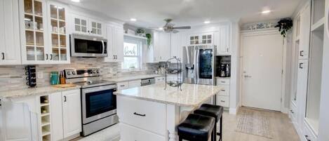 A modern kitchen with white cabinetry, a central island with stools, stainless steel appliances, and a ceiling fan.