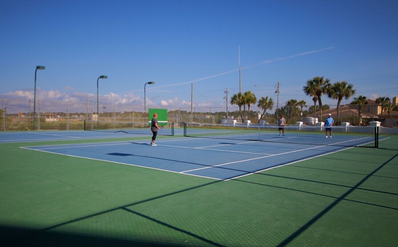 An outdoor tennis court with a green and blue surface, surrounded by palm trees and a clear sky.