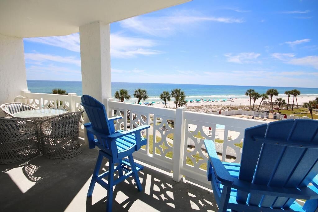 A balcony with blue chairs and a table overlooking a beach with palm trees and umbrellas.