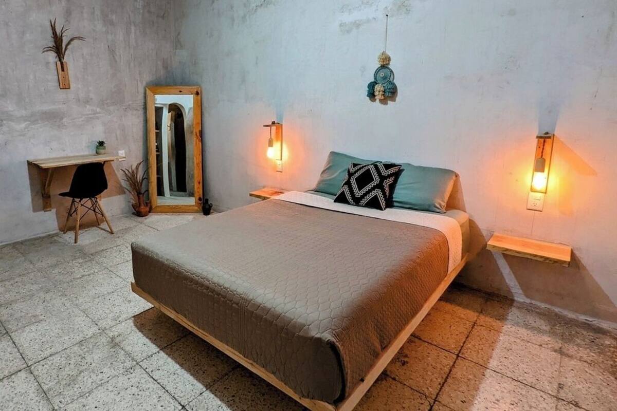 Une chambre à coucher avec un grand lit, une table de chevet en bois, un petit bureau et deux lampes fixées au mur.