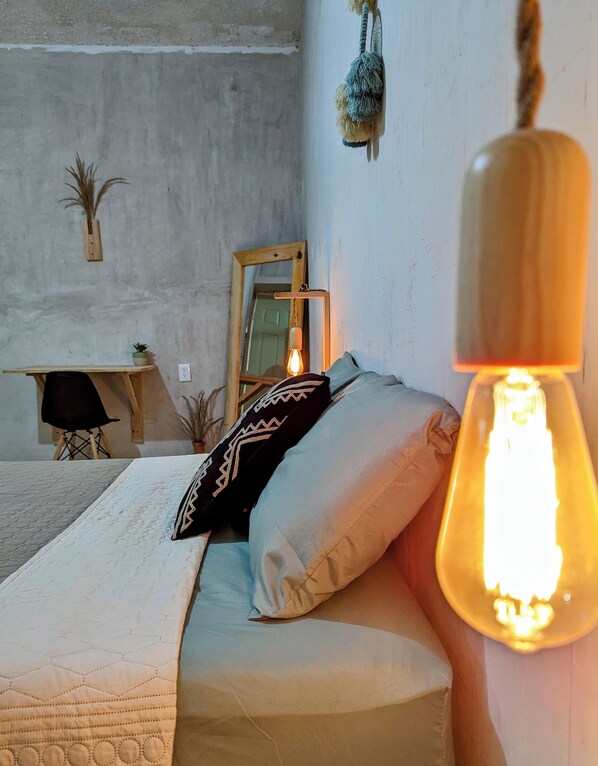 A bedroom with a bed, pillows, a hanging light fixture, a mirror, and a small table with a plant.