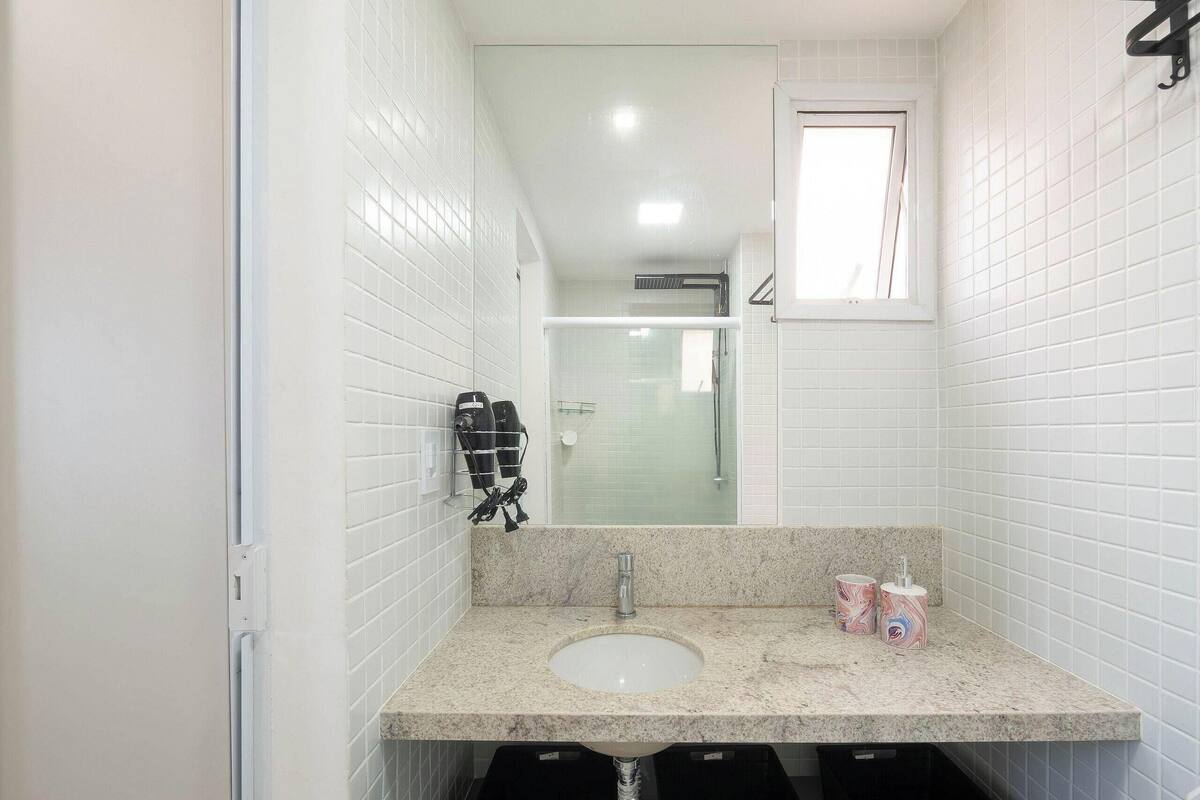 Une salle de bain avec des murs recouverts de carreaux blancs, un comptoir en granit, un lavabo, un miroir et une fenêtre.