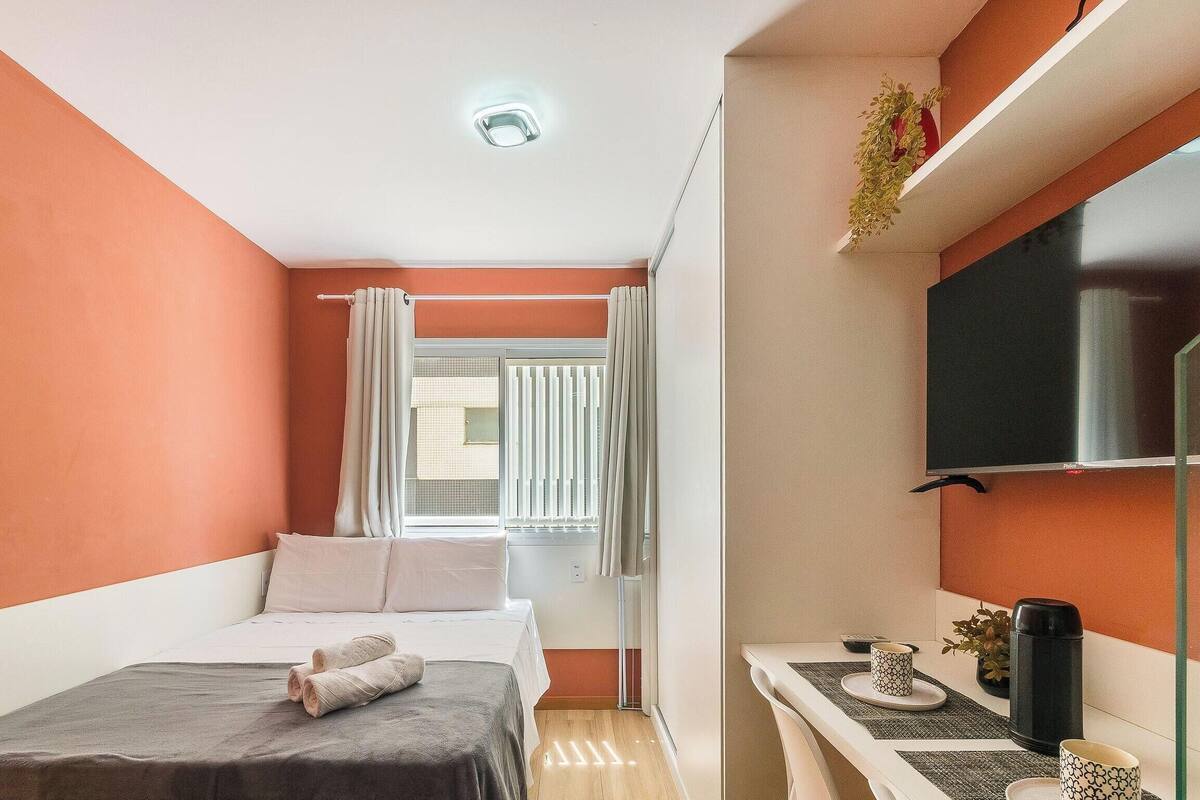 Une chambre d’hôtel de taille réduite, comprenant un lit, une petite table avec une plante, un téléviseur et une fenêtre avec des rideaux.