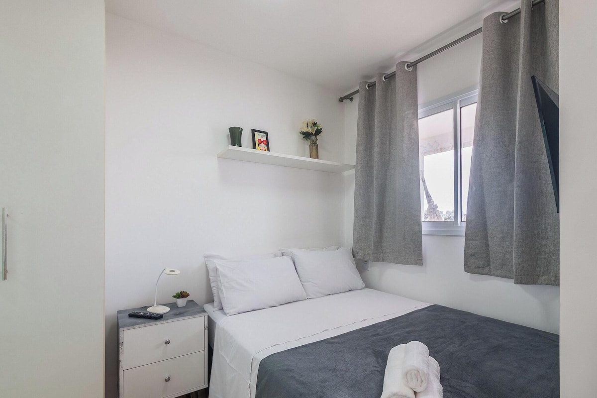 Une chambre à coucher comprenant un lit, une table de chevet, une commode, une fenêtre avec des rideaux et une étagère ornée d’objets décoratifs.