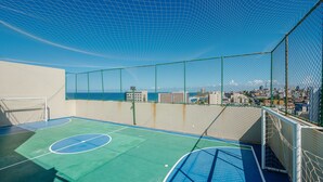 An outdoor basketball court with a netted enclosure, a goalpost, and a view of a cityscape and ocean. - Condo. w/ Pool and Gym 70 m From the Beach - SMA (Salvador)