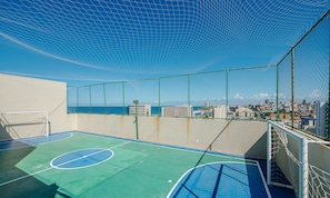 An outdoor basketball court with a netted enclosure, a goalpost, and a view of a cityscape and ocean. - Condo. w/ Pool and Gym 70 m From the Beach - SMA (Salvador)