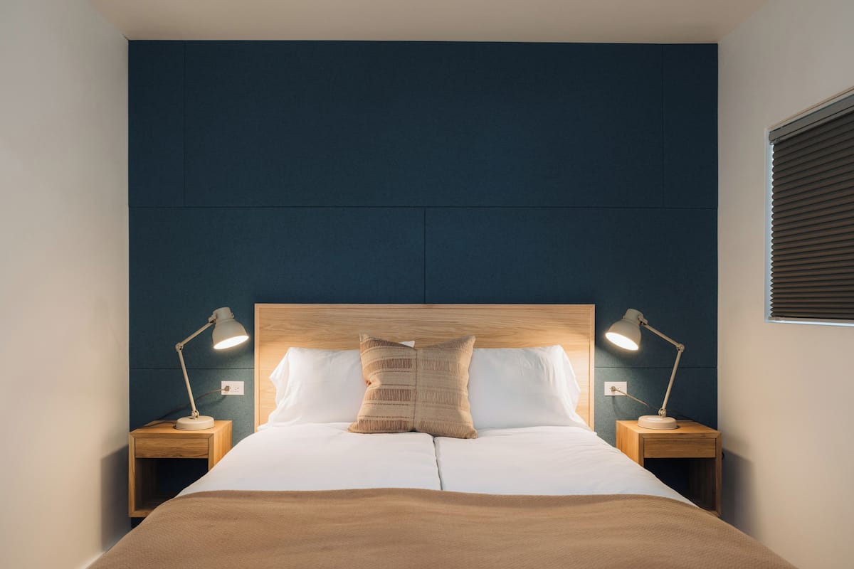 A neatly made bed with two bedside lamps, a wooden headboard, and a dark wall behind.