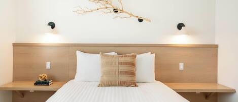 A hotel room with a wooden headboard, two bedside tables, a bed with white bedding, a textured throw blanket, and wall-mounted lighting.