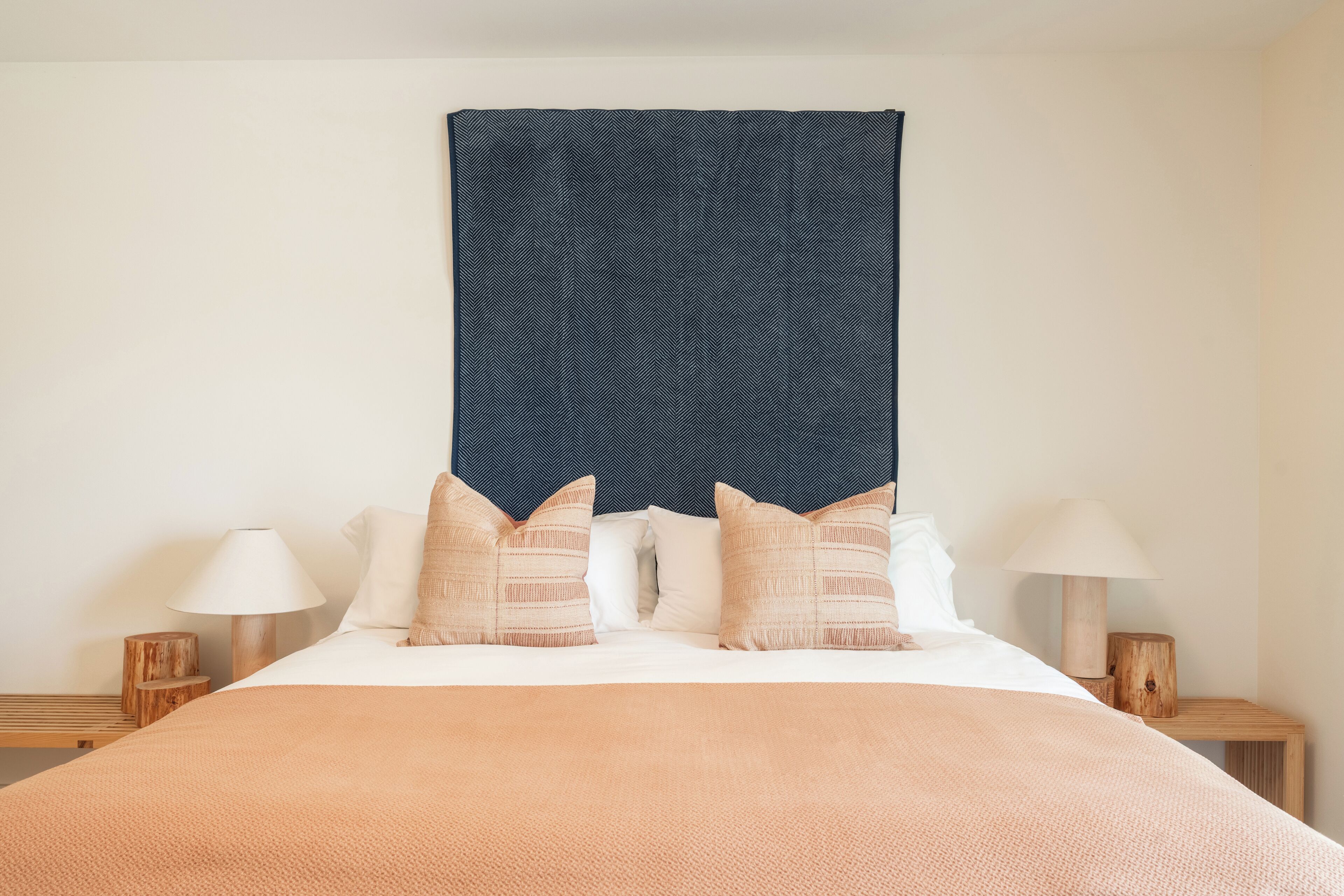 A neatly made bed with a peach-colored blanket, two beige pillows, and a dark blue patterned headboard. There are two bedside tables with lamps on either side of the bed.