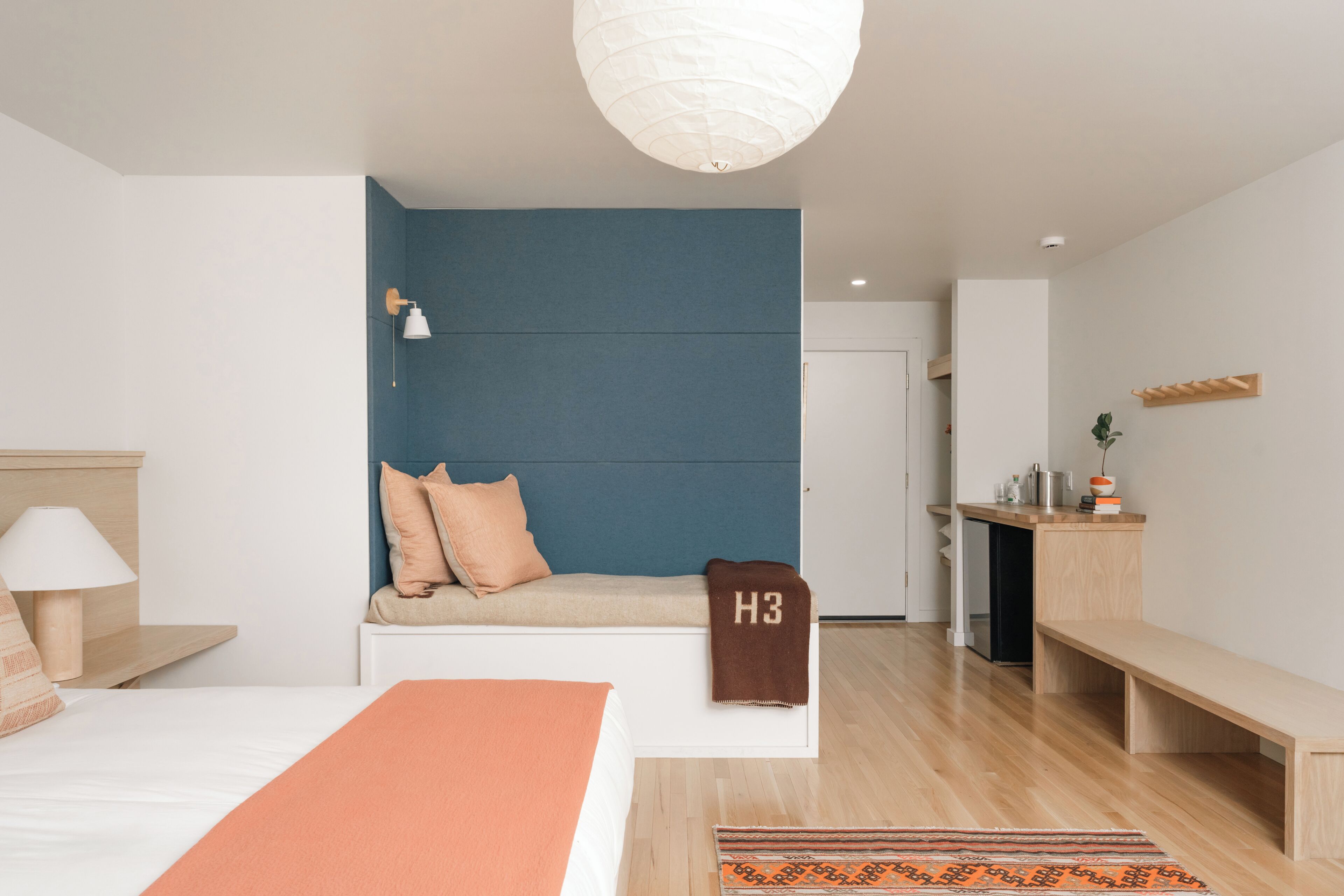 A modern hotel room with a bed, bedside table, lamp, bench, and a small kitchenette area.