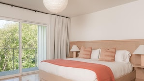 A bedroom with a large bed, two bedside lamps, a view of trees through a sliding door, and a patterned rug. - Mendocino Cove (Fort Bragg)