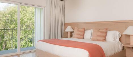 A bedroom with a large bed, two bedside lamps, a view of trees through a sliding door, and a patterned rug.