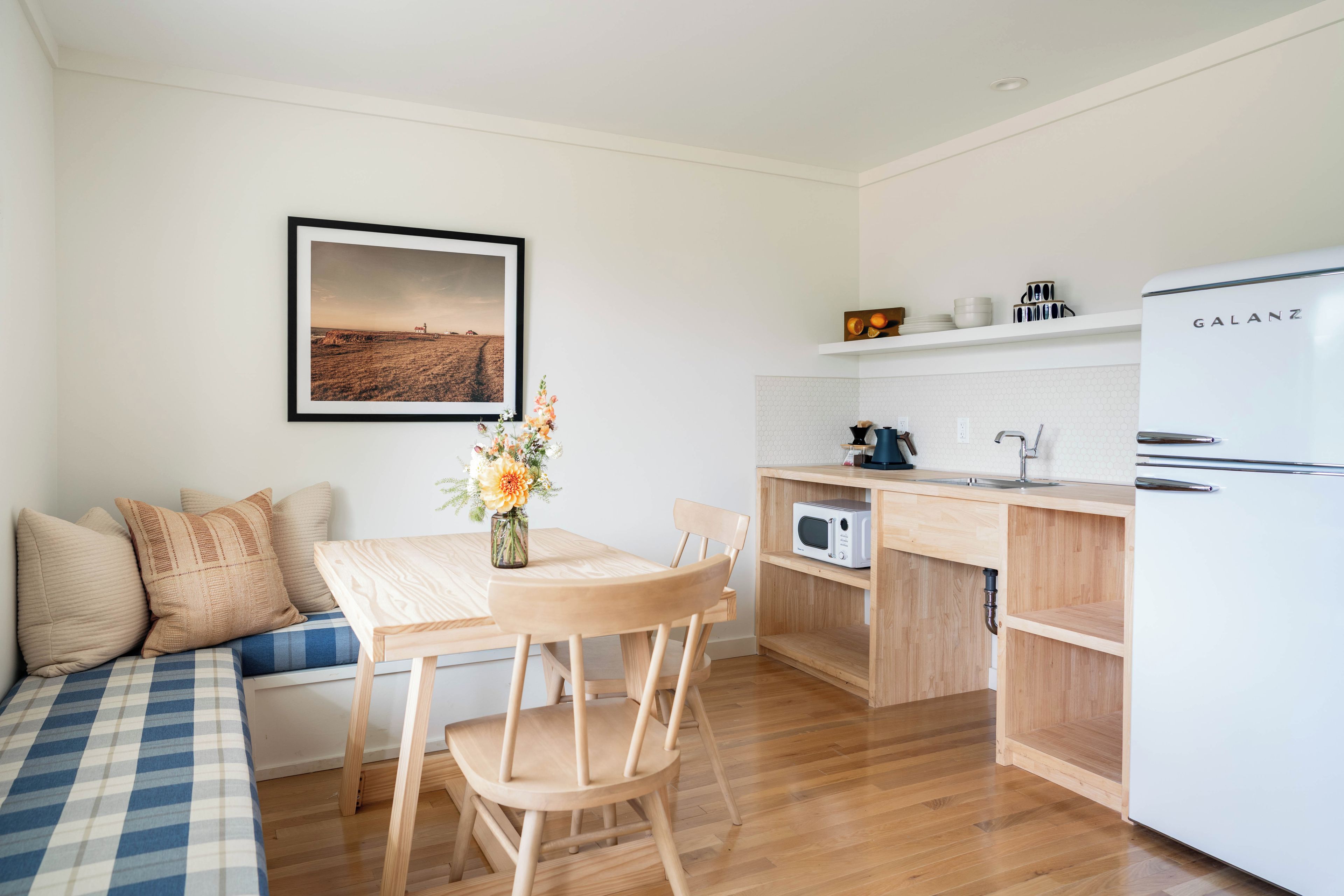 Una cocina compacta con microondas integrado, un refrigerador blanco Galanz, una mesa de comedor de madera con sillas, una pequeña planta en maceta y una fotografía enmarcadas de un paisaje en la pared.