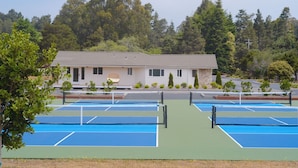 A tennis court complex with multiple courts, a building in the background, and surrounding trees. - Mendocino Cove (Fort Bragg)