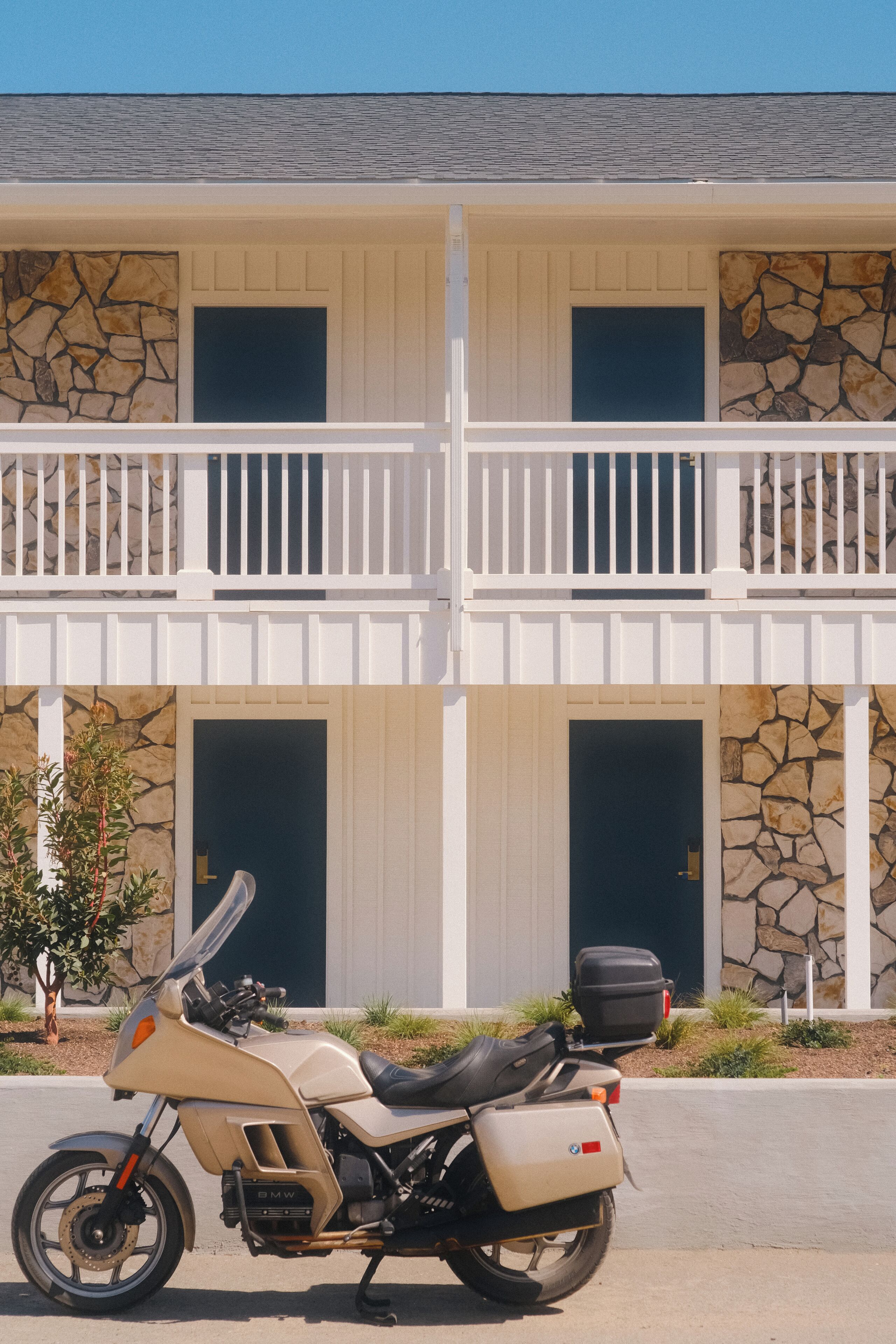 A two-story building with a stone facade, a balcony with white railings, and a parked motorcycle in front.